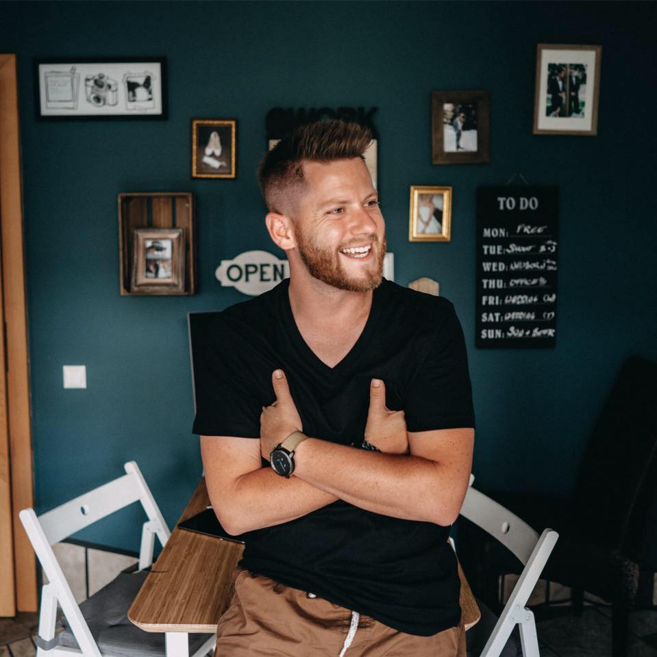 Ein lächelnder Mann mit kurzen Haaren und einem Bart sitzt mit verschränkten Armen auf einem Tisch und trägt ein schwarzes T-Shirt und braune Shorts. Hinter ihm befindet sich eine dunkle, blaugrüne Wand mit gerahmten Fotos, einer To-do-Tafel und weißen Stühlen.