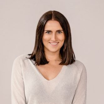 Eine Frau mit glattem braunem Haar, bekleidet mit einem hellgrauen Pullover, locker sitzenden Bluejeans und Turnschuhen, steht auf einem Bein und lächelt in die Kamera vor einem schlichten hellgrauen Hintergrund.