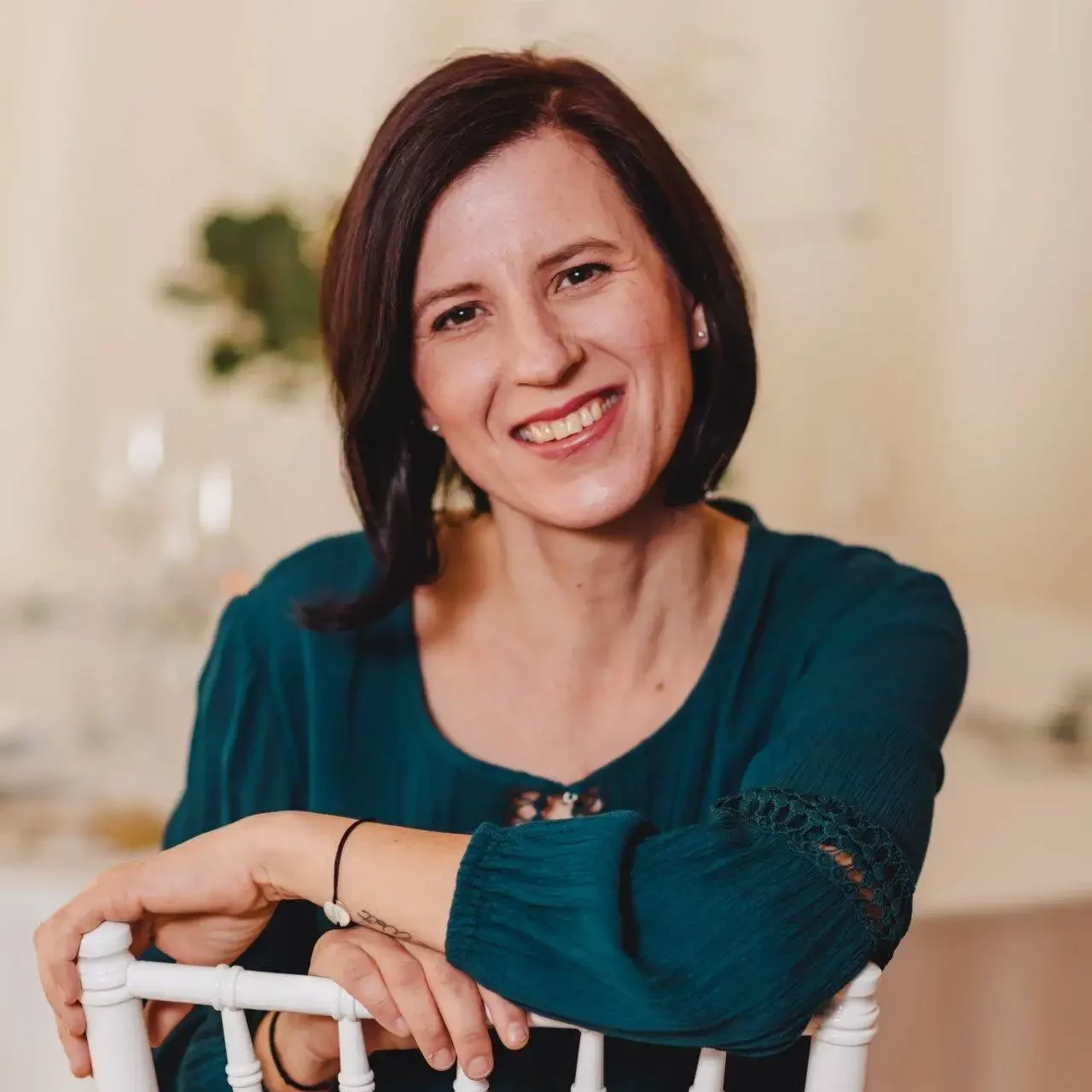 Eine Frau mit kurzen braunen Haaren und einem blaugrünen Kleid sitzt auf einem weißen Stuhl und lächelt warm in die Kamera. Der Hintergrund ist sanft verschwommen, ein Tisch und etwas Grün sind zu sehen.