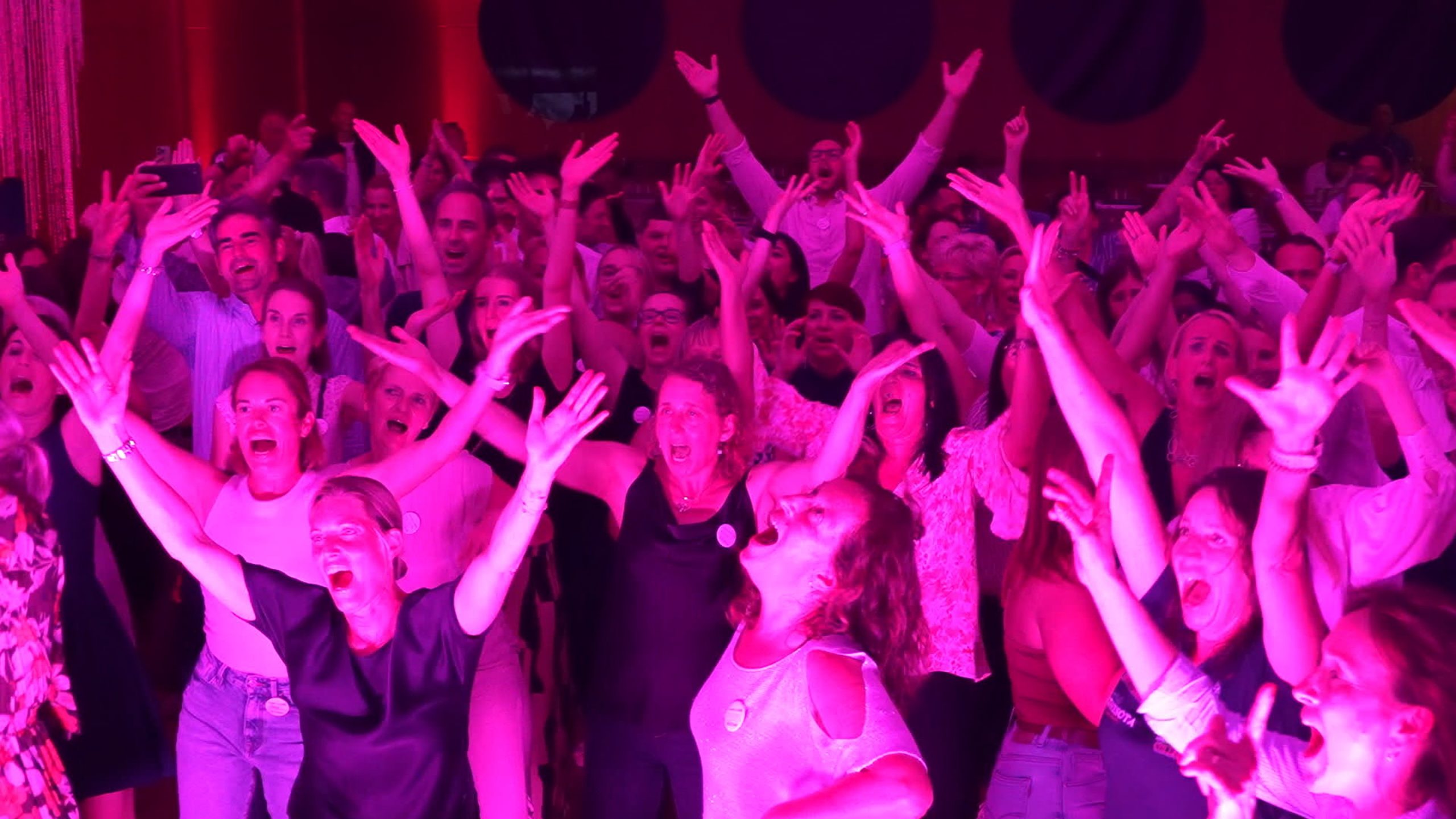 Eine große Gruppe von Menschen steht bei einer Indoor-Veranstaltung dicht beieinander, hebt aufgeregt die Arme und jubelt unter pulsierendem rosa und lila Licht - perfekt für alle, die hochzeit planen mit lebendiger DJ Musik.