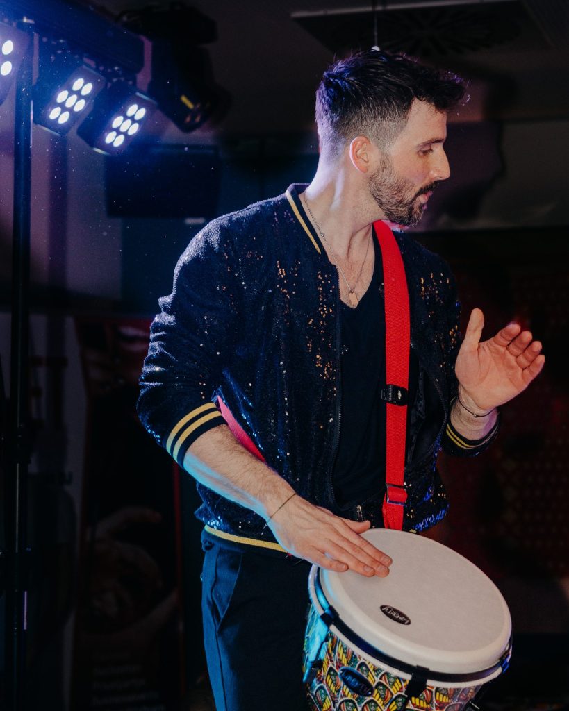 Ein Mann mit gestutztem Bart spielt im Scheinwerferlicht auf einer bunten Handtrommel, trägt eine dunkle Jacke mit Pailletten und einen roten Gurt - ein energiegeladener Live-Act für hochzeit österreich, bei dem eine Hand erhoben ist, als würde er den Rhythmus halten.