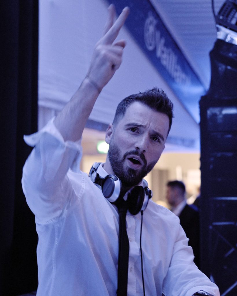 Ein Mann mit Kopfhörern um den Hals, der ein weißes Hemd und eine schwarze Krawatte trägt, gestikuliert mit zwei erhobenen Fingern, während er in einer blau beleuchteten Halle neben einem DJ-Equipment steht - perfekt als Ihr DJ für Weihnachtsfeiern.