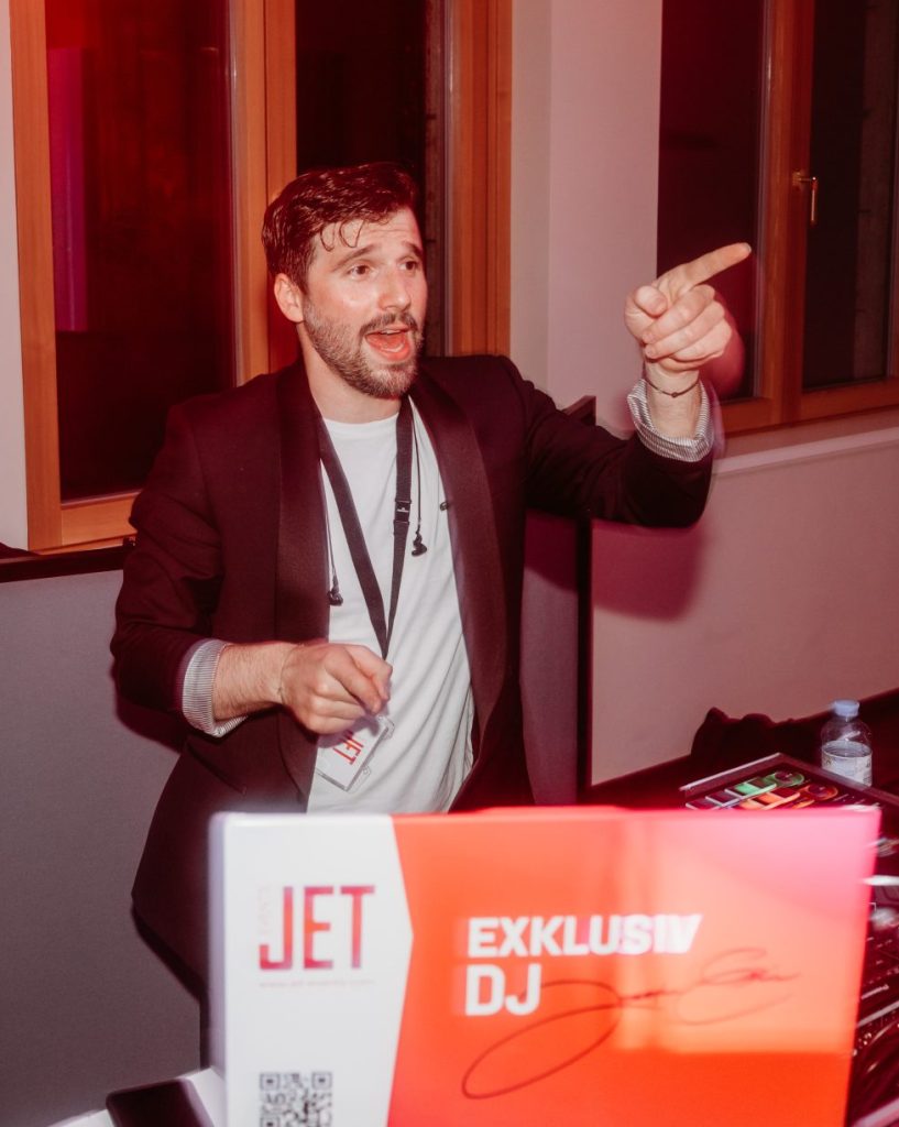 Ein DJ in einem dunklen Blazer, möglicherweise Joseph Erin, steht hinter einem Pult mit der Aufschrift "JET EXKLUSIV DJ", gestikuliert und lächelt. Der lebendige Raum mit roter Beleuchtung und großen Fenstern deutet auf einen energiegeladenen hochzeits-dj oder live act für hochzeit Österreich hin.