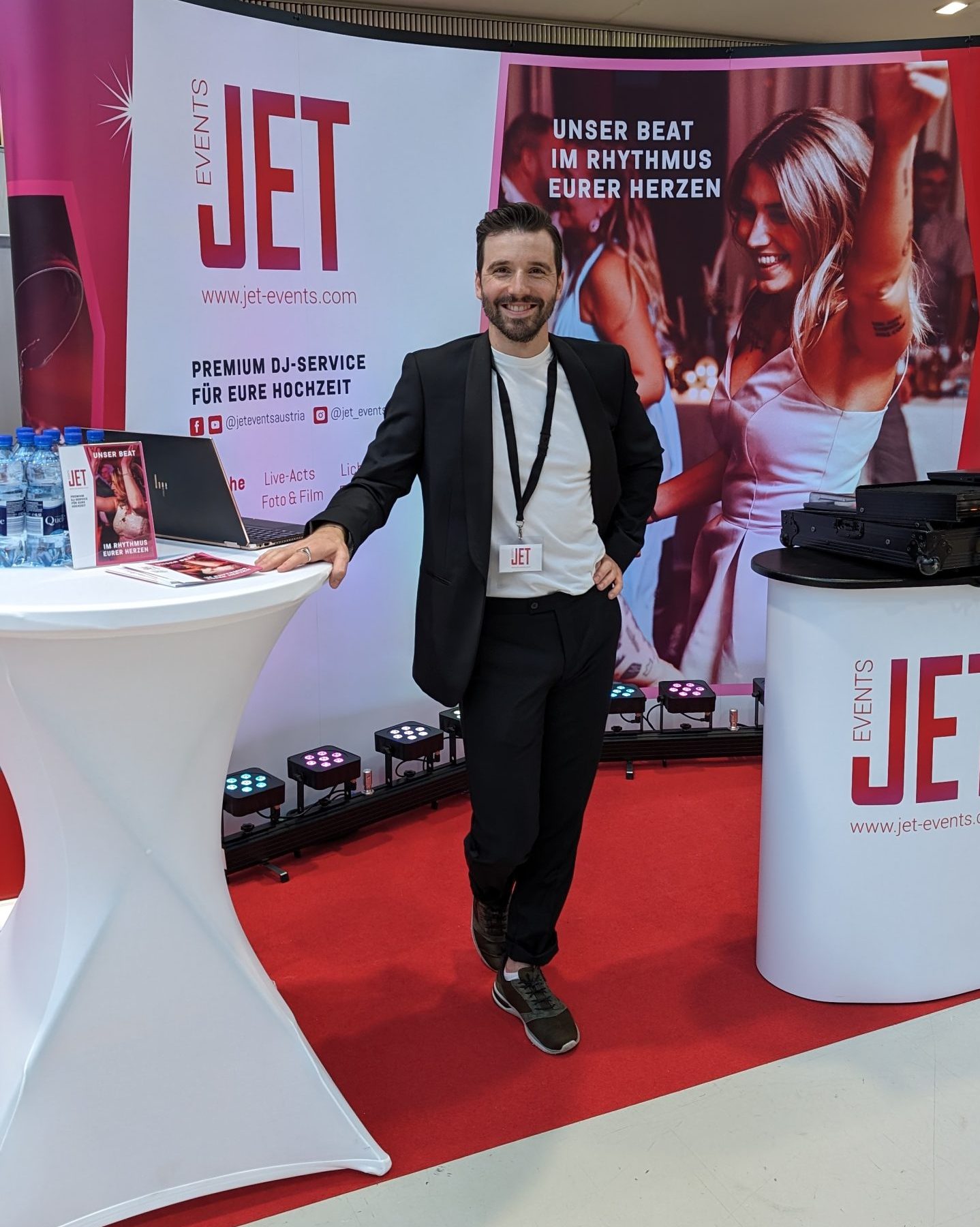 Ein lächelnder Mann im Anzug steht am Stand von JET Events auf der Hochzeitsmesse Oberösterreich im Design Center in Linz. Er wirbt mit Bannern für Premium-DJ-Services für Hochzeiten und zeigt ein lebhaftes Foto von tanzenden Menschen im Hintergrund.