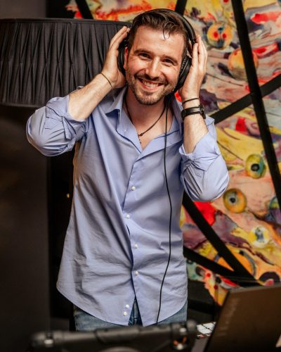 Ein lächelnder Mann in einem hellblauen Hemd steht in einem Haus und hält sich Kopfhörer an die Ohren, während er Tracks wie ein echter hochzeits dj wien mischt. Vor ihm stehen DJ-Equipment und ein Laptop, im Hintergrund bunte abstrakte Kunst und gelbe Vorhänge.