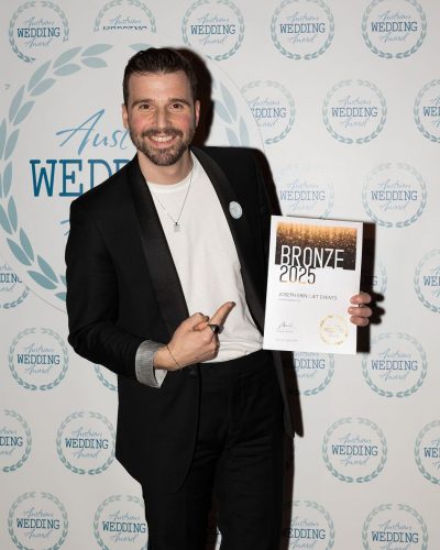 Ein lächelnder Mann in schwarzem Anzug und weißem Hemd hält eine "Bronze 2025"-Auszeichnungsurkunde in der Hand und zeigt auf sie. Er steht vor einer Kulisse mit "Wedding"-Logos im Step-and-Repeat-Verfahren, die hochzeits-dj Joseph Erin bei der Veranstaltung perfekt einfängt.