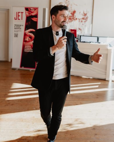 Ein Mann in einem schwarzen Anzug hält ein Mikrofon in der Hand und spricht, während er mit der Hand gestikuliert, in einem hellen, modernen Raum mit Holzboden und zeitgenössischer Kunst im Hintergrund.