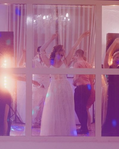 Eine Gruppe von Menschen, darunter eine Braut in einem weißen Kleid, tanzt freudig hinter großen Fensterscheiben auf einem scheinbaren Hochzeitsempfang. Die Szene ist warm beleuchtet und lebendig, mit erhobenen Armen und sichtbarem Lächeln.