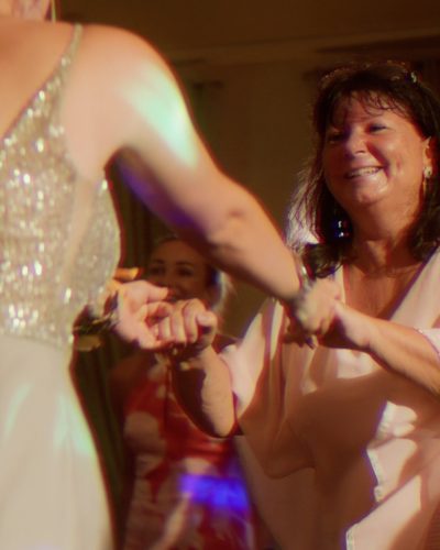 Zwei Frauen lächeln und halten sich an den Händen, während sie auf einer lebhaften Indoor-Veranstaltung tanzen. Die eine trägt ein glitzerndes Kleid mit offenem Rücken, die andere ein helles Outfit. Auch die Menschen im Hintergrund genießen die Feier.