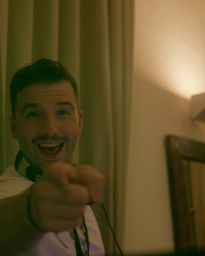 Ein Mann mit Kopfhörern um den Hals lächelt und zeigt in die Kamera. Er steht in einem Innenraum vor beigen Vorhängen und einem Spiegel mit warmer Beleuchtung.