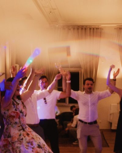 Eine Gruppe formell gekleideter Menschen tanzt fröhlich in einem Innenraum unter sanften, farbenfrohen Lichtern, hebt die Arme und winkt mit Leuchtstäben zur Feier. Die fröhliche Atmosphäre erinnert an eine Hochzeit oder Party.