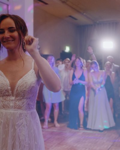 Eine Braut in einem weißen Spitzenkleid hält einen Blumenstrauß und lächelt, während sie sich darauf vorbereitet, ihn einer Gruppe von Frauen zuzuwerfen, die hinter ihr auf einer Hochzeitsfeier mit bunten Lichtern warten.