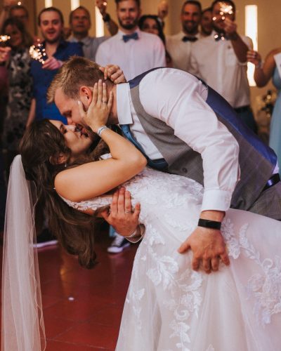 Ein Bräutigam küsst seine Braut auf der Tanzfläche, umgeben von lächelnden Gästen in formeller Kleidung mit Wunderkerzen. Die Braut trägt ein weißes Kleid und einen Schleier; der Bräutigam trägt eine Weste und eine Krawatte. | Hochzeits-DJ