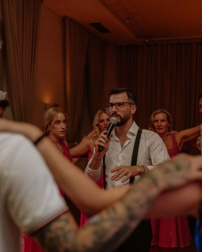 Ein hochzeits DJ Kärnten spricht mit einem Mikrofon zu einer Gruppe, die in einem warm beleuchteten Raum mit beigen Vorhängen im Kreis steht und die Arme umeinander legt. Einige TeilnehmerInnen tragen festliche Outfits und Accessoires.