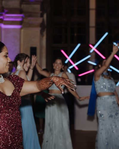 Eine Frau in einem roten Paillettenkleid mit kunstvollem Henna auf den Armen tanzt lächelnd, während ein hochzeits DJ Wien die Menge anheizt und andere Frauen in blauen und blaugrünen Outfits sich fröhlich unter Neonlichtern bewegen.