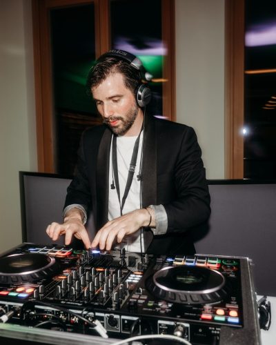 Ein hochzeits DJ Wien mit Kopfhörern und schwarzem Anzug mischt bei einer Indoor-Veranstaltung Musik an einem professionellen DJ-Controller, während im Hintergrund bunte Lichter reflektieren und eine Wasserflasche an der Seite steht.