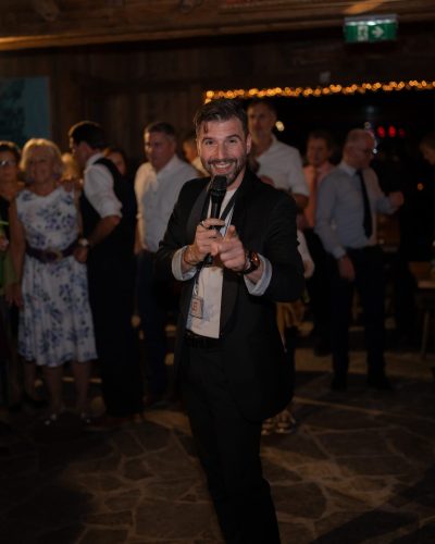 Ein Mann im Anzug, der ein Mikrofon in der Hand hält und lächelnd in die Kamera zeigt, während im Hintergrund eine Gruppe versammelt ist – eine ideale Szene für jeden DJ für Hochzeiten und DJ für Veranstaltungen, der die Feier anheizen möchte.