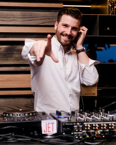 Ein lächelnder Mann in einem weißen Hemd steht hinter einem DJ-Equipment, trägt Kopfhörer und zeigt in einer belebten Innenumgebung in Richtung Kamera.