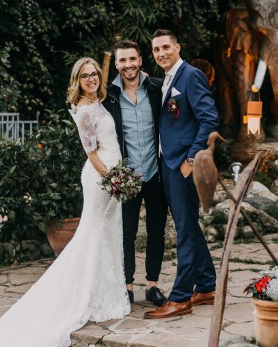 Eine Braut in einem weißen Kleid und ein Bräutigam in einem blauen Anzug posieren mit einem Gast im Freien auf dem Kleinsasserhof in Spittal an der Drau, umgeben von Grün und Blumen, mit einer großen Elefantenstatue im Hintergrund. Alle lächeln bei der Hochzeit.